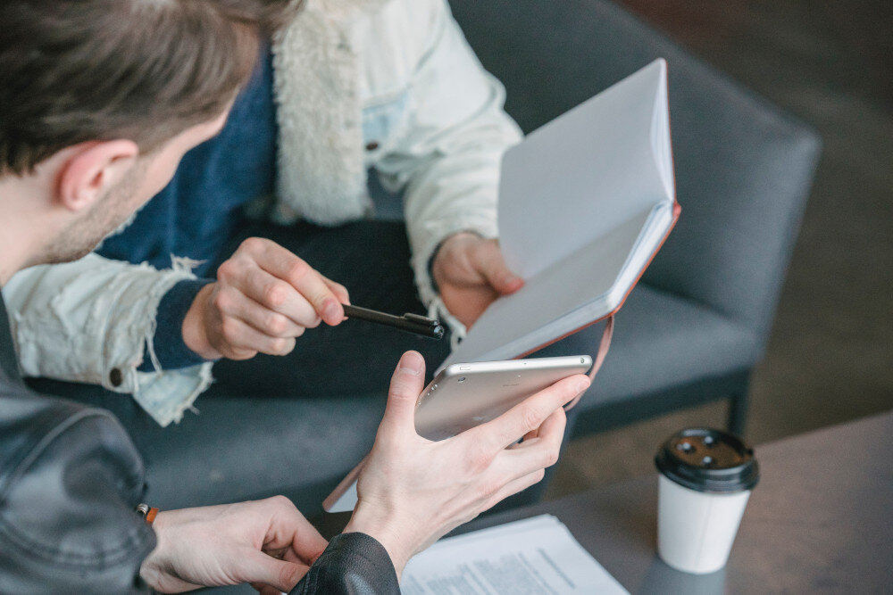 Zwei Kollegen sitzen auf bequemen Sitzmöbeln und besprechen einen Business Case. Ein Kollege hält dabei ein Tablet, der andere ein Notizbuch. | © pexels-laura-tancredi-7083913